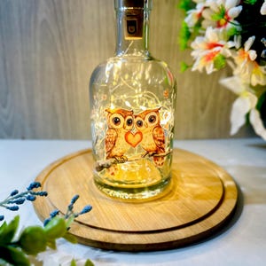May include: A clear glass bottle with a cork stopper, lit by fairy lights. The bottle has a decorative image of two owls with a heart. The bottle sits on a round wooden tray. Flowers are visible in the background.