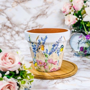 May include: A white ceramic plant pot featuring a painted design of a wooden fence, birds, pink flowers, and blue flowers. The pot is on a round wooden tray. The background is a marble-patterned surface.