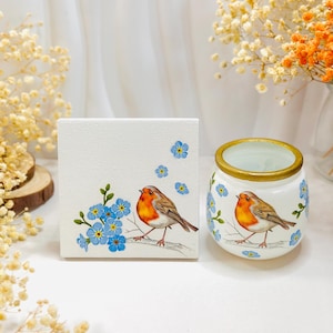 May include: A square white canvas and a small white ceramic jar, both featuring a hand-painted illustration of a robin perched on a branch surrounded by blue forget-me-not flowers. The jar has a gold-painted rim and a rounded shape.