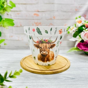 May include: White ceramic plant pot with a Highland cow design, adorned with a floral crown. The pot's rim is decorated with floral and leaf motifs. It rests on a round wooden coaster, suitable for indoor or outdoor use.