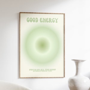 May include: A framed print with the words "GOOD ENERGY" in light green, above a green gradient circle. Below the circle, the text reads "FOCUS ON ALL THE GOOD SURROUNDING YOU". The print is in a room with a white chair and a wooden table.