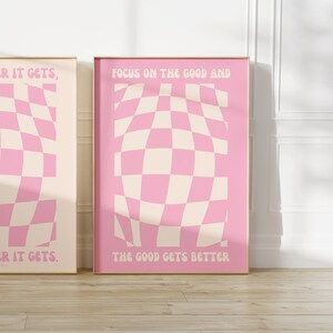 May include: Two pink and cream checkered posters with motivational quotes. The left poster says "_R IT GETS, ER IT GETS." The right poster says "FOCUS ON THE GOOD AND THE GOOD GETS BETTER."