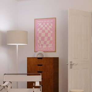 May include: A pink and white checkered poster with the text "Focus on the good and the good gets better". The poster is framed in wood and hanging on a white wall.