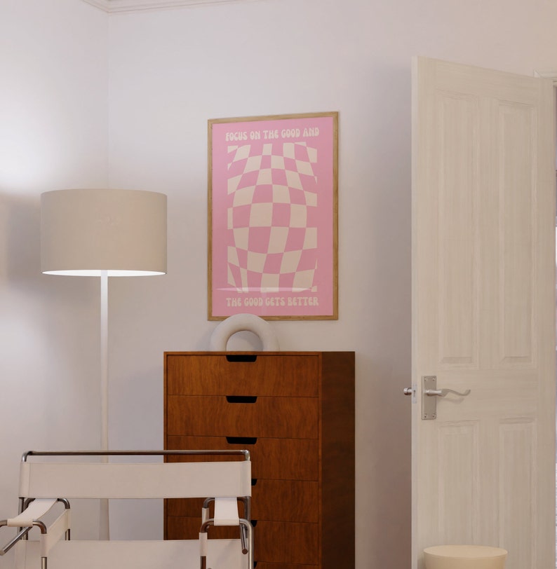 May include: A pink and white checkered poster with the text "Focus on the good and the good gets better". The poster is framed in wood and hanging on a white wall.