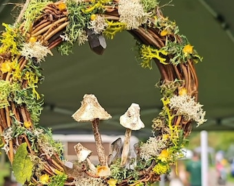 以下が含まれることがあります： 素朴な茶色の小枝の土台に、苔、小さな黄色の花、白い陶器のキノコ3つが飾られた手作りリース。リースは紐で吊り下げられており、3つのクリスタルが垂れ下がっています。