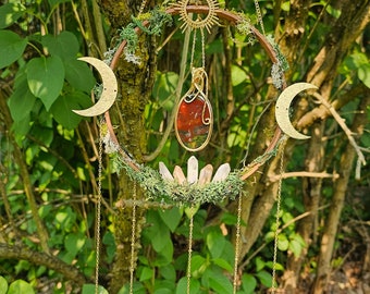 May include: A gold metal dream catcher with a wooden hoop, moss, crystals, and a red and green stone pendant. The dream catcher is hanging from a tree branch.