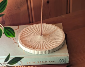 May include: White ceramic incense holder with a fluted design and speckled texture. A brown incense stick is standing upright in the center of the holder.