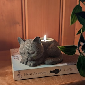 May include: A grey cat-shaped candle holder with a lit candle. The cat rests on a book titled "The Traveler" by Hiro Arikawa. The candle holder is made of a textured material. The scene is set on a wooden surface.