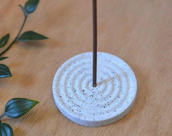 May include: White ceramic incense holder with a textured, circular design. A brown incense stick is placed in the center hole.