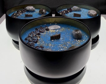 Puede incluir: Tres velas negras en forma de cuenco con cera azul, decoradas con piedras, cuentas oscuras y una mecha de madera. Las velas tienen un borde dorado y se asientan sobre una base negra. La cera está salpicada de purpurina dorada.