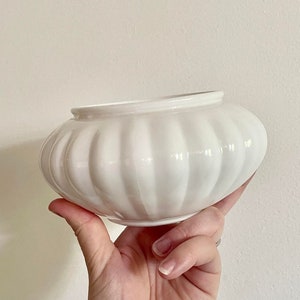 May include: A white, ribbed ceramic bowl with a rounded shape. The bowl is held up by a hand, showcasing its smooth, glossy surface. The design features vertical ridges, adding texture to the otherwise simple, elegant piece. A decorative item.