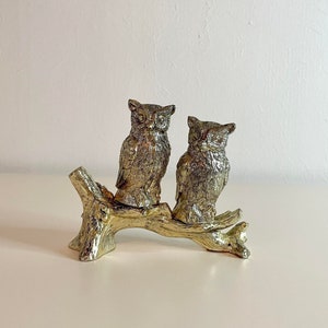 May include: Two silver owls perched on a branch, facing forward. The owls have detailed feathers and large, round eyes. The branch is made of metal and has a rustic finish.