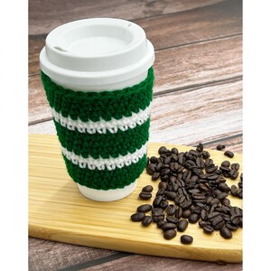 May include: A white ceramic travel mug with a white lid and a green and white striped crochet cozy. The mug sits on a wooden cutting board next to a pile of roasted coffee beans. The background is a wooden surface.