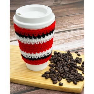 Op de afbeelding: Een witte keramische reismok met een wit deksel en een rood, zwart en wit gehaakte hoes. De mok staat op een houten snijplank naast een stapel koffiebonen. De hoes is gemaakt van dik garen.