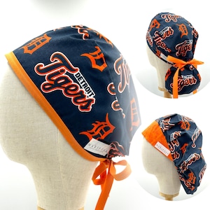 May include: A blue and orange surgical scrub hat with a repeating pattern of the Detroit Tigers logo. The hat has a tie on the back.