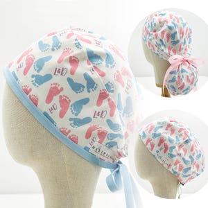 Puede incluir: Un gorro de quirófano blanco con un patrón de huellas de pies de bebé azul y rosa. El gorro tiene un lazo azul y el texto "L&D" se repite en todo el patrón. El gorro también tiene una pequeña etiqueta blanca con el texto "MARIPOSA" en ella.
