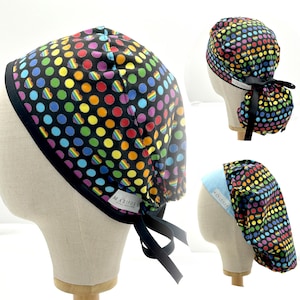 Op de afbeelding: Drie zwarte scrubmutsen met regenboog stippen en zwarte banden. De mutsen hebben een blauw en wit label met de tekst "Mariposa" erop.