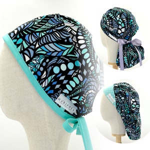 Può includere: Un cappello chirurgico blu e nero con motivo, con bordo e cravatta turchese. Il cappello ha un'etichetta che recita "Mariposa Natural Textiles Made in USA".