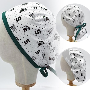 May include: White and green surgical scrub hat with a repeating pattern of the Michigan State Spartan logo and the letter "S". The hat has a green elastic band and green ties.