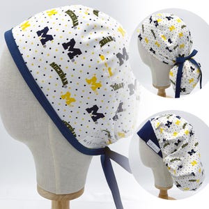 May include: A white scrub cap with a navy blue trim and ties. The cap is patterned with navy blue and yellow "M" logos and the word "Michigan" in navy blue. The background has small black and yellow polka dots.