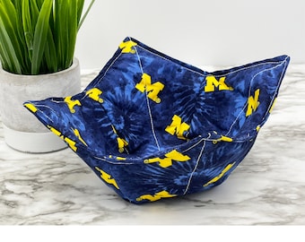 May include: A fabric bowl cozy featuring a dark blue tie-dye pattern with repeating yellow block letter M logos. The square-shaped holder has four folded corners and white topstitching, designed to hold a bowl securely while protecting hands.