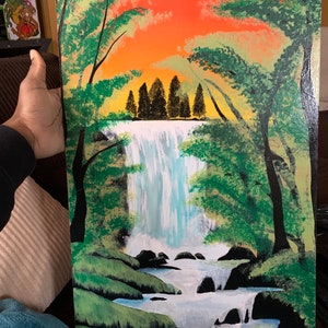Pode incluir: Uma pintura retangular numa superf&iacute;cie plana que retrata uma cascata a cair sobre rochas escuras para um riacho. A cena &eacute; emoldurada por &aacute;rvores verdes em ambos os lados, contra um c&eacute;u vibrante de p&ocirc;r do sol laranja e amarelo.