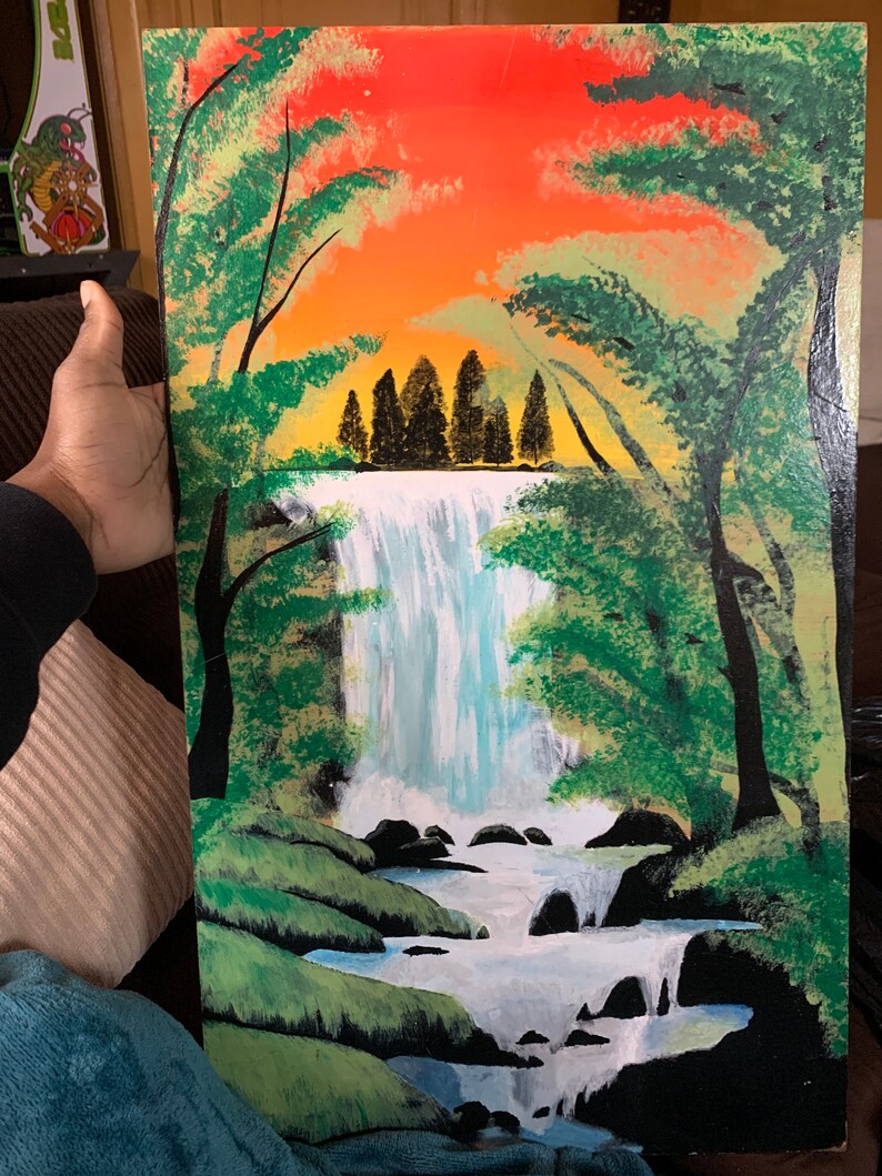 Pode incluir: Uma pintura retangular numa superf&iacute;cie plana que retrata uma cascata a cair sobre rochas escuras para um riacho. A cena &eacute; emoldurada por &aacute;rvores verdes em ambos os lados, contra um c&eacute;u vibrante de p&ocirc;r do sol laranja e amarelo.