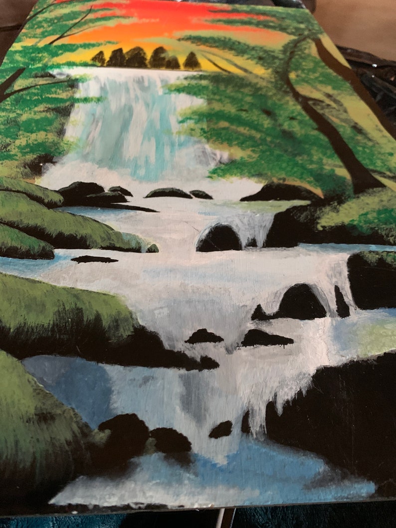 Pode incluir: Uma pintura de paisagem em tela retratando uma cachoeira fluindo sobre rochas escuras em um riacho. A cena apresenta folhagem verde vibrante, &aacute;rvores nas margens e um c&eacute;u de p&ocirc;r do sol laranja e amarelo brilhante no topo da composi&ccedil;&atilde;o.