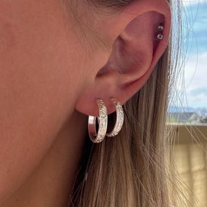 May include: A close-up of a person's ear with two silver hoop earrings. The earrings have a detailed floral pattern. The person's hair is long and light brown, and the background is a blurred view of the sky.
