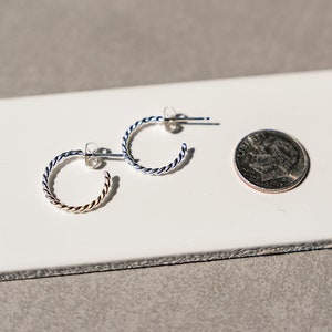May include: Two pairs of silver hoop earrings with a twisted design. The earrings are shown on a white tile surface next to a US dime for scale.