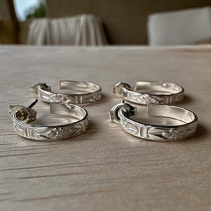 May include: Four silver hoop earrings with floral and geometric patterns. The earrings are displayed on a light-coloured wooden surface. Each earring has a post for pierced ears.