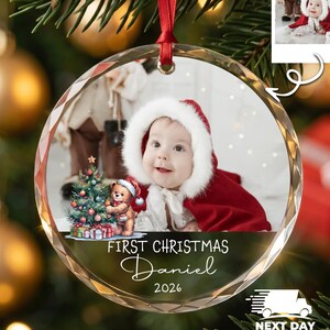 K&ouml;nnte beinhalten: Ein runder, klarer Weihnachtsbaumschmuck aus Glas mit einem Foto eines Babys in einem roten Kapuzenumhang mit wei&szlig;em Fellbesatz. Das Ornament zeigt eine Grafik eines Teddyb&auml;ren mit Weihnachtsbaum und den Text First Christmas Daniel 2026.