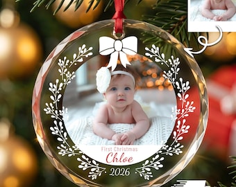 May include: A round, clear glass Christmas ornament featuring a printed photograph of a baby wearing a white flower headband. The ornament is decorated with a white bow and leaf pattern, with the text First Christmas, Chloe, and 2026 printed below.