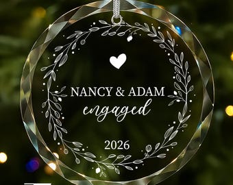 May include: A circular clear glass ornament with faceted edges, featuring a white engraved botanical wreath design. The center displays a heart icon, the text NANCY & ADAM, the word engaged, and the year 2026. It hangs from a silver cord.