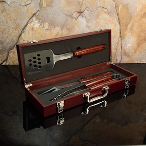 May include: A wooden box with a brown stain and a silver latch. The box contains a set of grilling tools with wooden handles. The tools are labeled "STEPHEN".