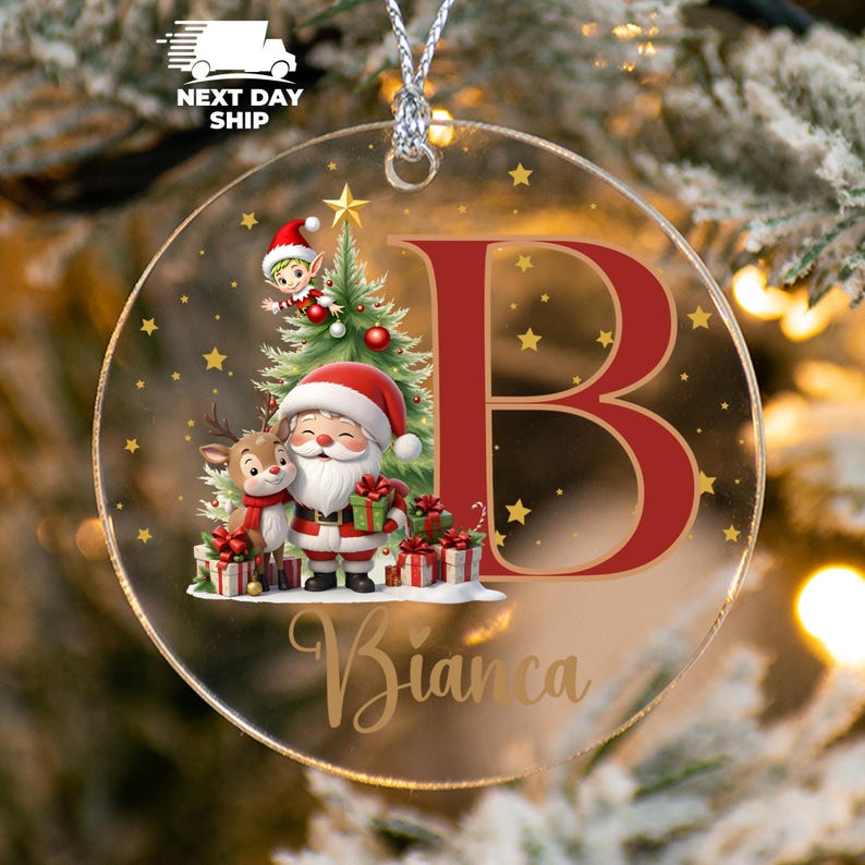 May include: A clear, round Christmas ornament with a festive design. It features Santa Claus, a reindeer, and an elf near a Christmas tree, with the letter "B" and the name "Bianca" in gold script. Gold stars and a silver string complete the design.