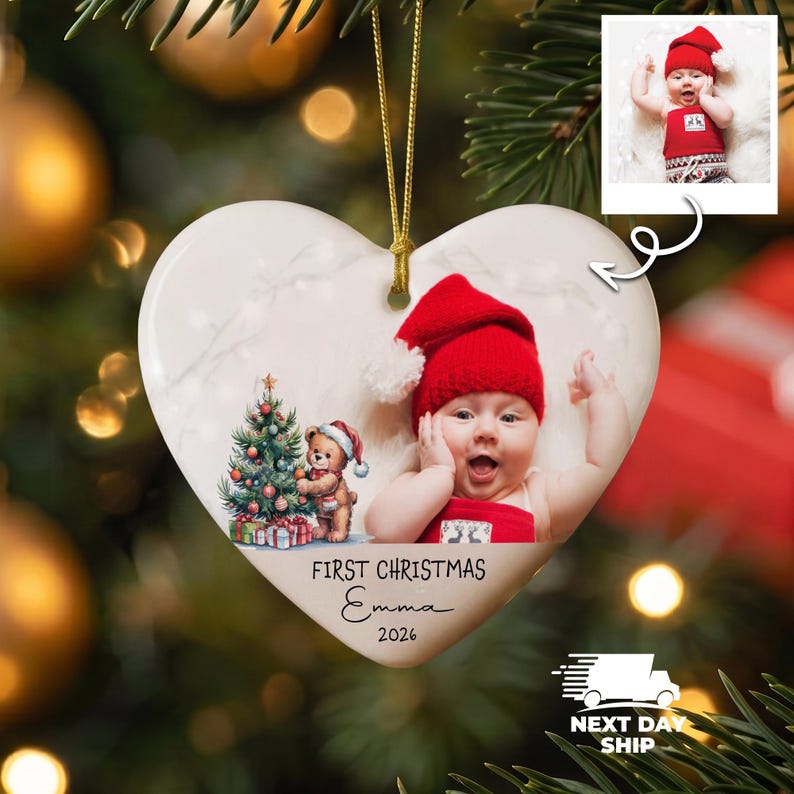 K&ouml;nnte beinhalten: Ein herzf&ouml;rmiger wei&szlig;er Keramikschmuck mit einem aufgedruckten Bild eines Babys mit roter M&uuml;tze und einem Teddyb&auml;ren neben einem Weihnachtsbaum. Der Text lautet FIRST CHRISTMAS Emma 2026. Eine goldene Schnur dient zum Aufh&auml;ngen am Baum.