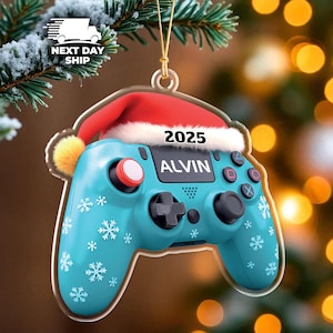 May include: A turquoise Christmas ornament shaped like a video game controller, adorned with snowflakes. It features a red Santa hat with the year 2025 and the name "ALVIN". The ornament is suspended by a gold string and set against a blurred background.