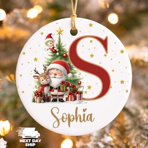 May include: A white ceramic Christmas ornament featuring Santa Claus, a reindeer, and an elf near a Christmas tree. The ornament has a red letter "S" and the name "Sophia" in gold script. Gold stars are scattered across the surface.