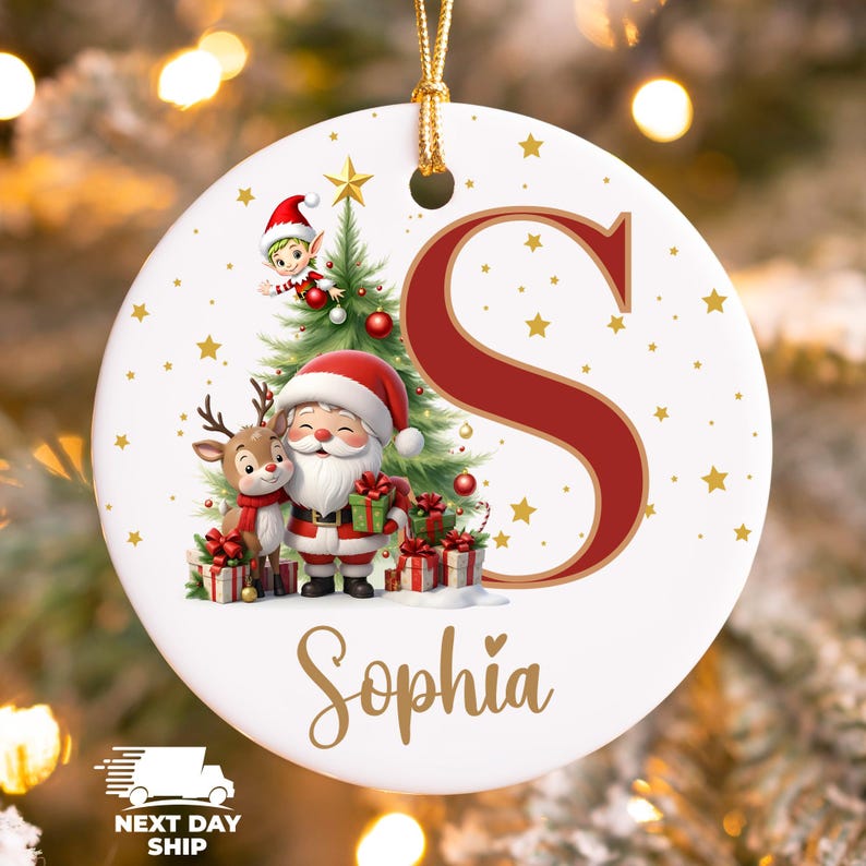 May include: A white ceramic Christmas ornament featuring Santa Claus, a reindeer, and an elf near a Christmas tree. The ornament has a red letter "S" and the name "Sophia" in gold script. Gold stars are scattered across the surface.