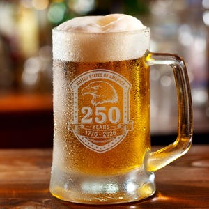 May include: A clear glass beer mug filled with golden beer and a thick white head. The mug has a handle and features an etched design with an eagle, the text "UNITED STATES OF AMERICA", and "250 YEARS 1776-2026".