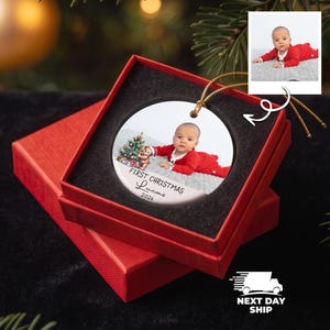 K&ouml;nnte beinhalten: Ein rundes, wei&szlig;es Keramikornament mit einem Foto eines Babys in roter Kleidung, das neben einem kleinen Weihnachtsbaum und einem Teddyb&auml;ren liegt. Der Text lautet First Christmas Lucas 2026. Das Ornament hat eine goldene Schnur und liegt in einer roten Box.