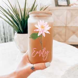 May include: A clear glass tumbler with a bamboo lid and a clear straw. The tumbler features a pink lotus flower decal and the name "Emily" in pink script. The tumbler is filled with a light brown liquid.