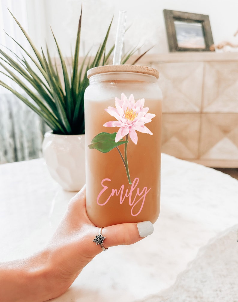 May include: A clear glass tumbler with a bamboo lid and a clear straw. The tumbler features a pink lotus flower decal and the name "Emily" in pink script. The tumbler is filled with a light brown liquid.