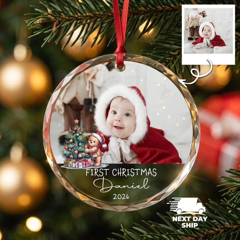 K&ouml;nnte beinhalten: Ein runder, klarer Weihnachtsbaumschmuck aus Glas mit einem Foto eines Babys in einem roten Kapuzenumhang mit wei&szlig;em Fellbesatz. Das Ornament zeigt eine Grafik eines Teddyb&auml;ren mit Weihnachtsbaum und den Text First Christmas Daniel 2026.