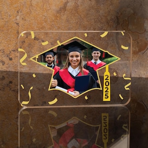 May include: A clear acrylic photo frame with a gold graduation cap design. The frame features a photo of three graduates wearing caps and gowns. The cap has a tassel with the year "2025" printed on it.