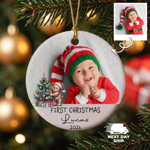 K&ouml;nnte beinhalten: Ein runder, wei&szlig;er Keramik-Weihnachtsbaumschmuck mit einem Foto eines Babys, das eine rot-wei&szlig; gestreifte M&uuml;tze mit gr&uuml;nem Rand tr&auml;gt. Das Ornament zeigt eine Grafik eines Teddyb&auml;ren neben einem Weihnachtsbaum und den Text First Christmas Lucas 2026.