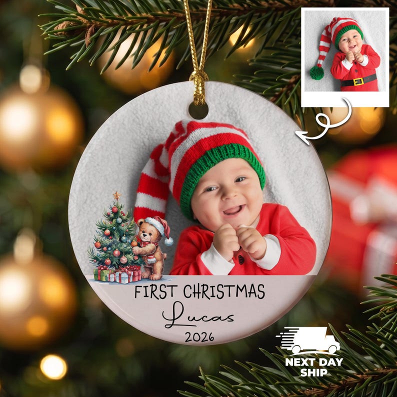 K&ouml;nnte beinhalten: Ein runder, wei&szlig;er Keramik-Weihnachtsbaumschmuck mit einem Foto eines Babys, das eine rot-wei&szlig; gestreifte M&uuml;tze mit gr&uuml;nem Rand tr&auml;gt. Das Ornament zeigt eine Grafik eines Teddyb&auml;ren neben einem Weihnachtsbaum und den Text First Christmas Lucas 2026.