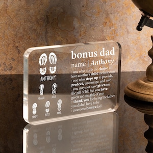 May include: A clear acrylic block with a white inscription that reads "bonus dad name | Anthony" and a definition of what a bonus dad is. The block also features a footprint design with the name "Anthony" and the names of other family members.