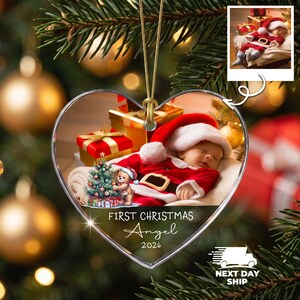 K&ouml;nnte beinhalten: Ein herzf&ouml;rmiger Weihnachtsbaumschmuck mit einem schlafenden Baby im Weihnachtsmannkost&uuml;m, einem kleinen Teddyb&auml;ren und einem geschm&uuml;ckten Baum. Der Text lautet FIRST CHRISTMAS Angel 2026. Der Schmuck h&auml;ngt an einer goldenen Kordel.